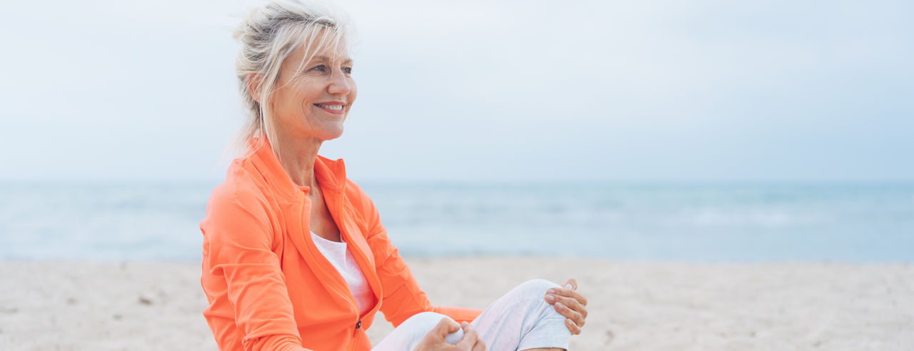 Happy supple mature blond woman seated in the golden sand on a beach looking to the side of the frame with a beaming smile against an ocean backdrop